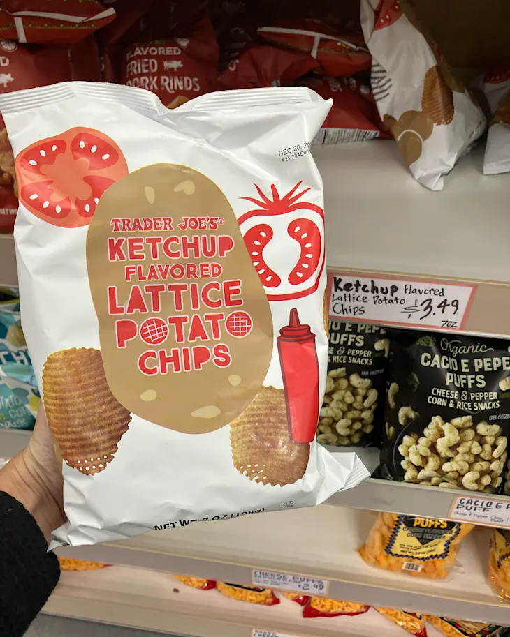 Ketchup flavored lattice potato chips in a white bag held in a grocery aisle, with other snacks in the background.