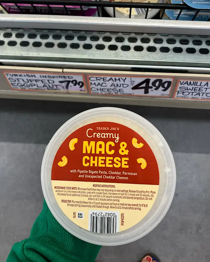 Creamy mac and cheese container from Trader Joe's held by a person wearing a green sweater, with price tags for other products in the background.