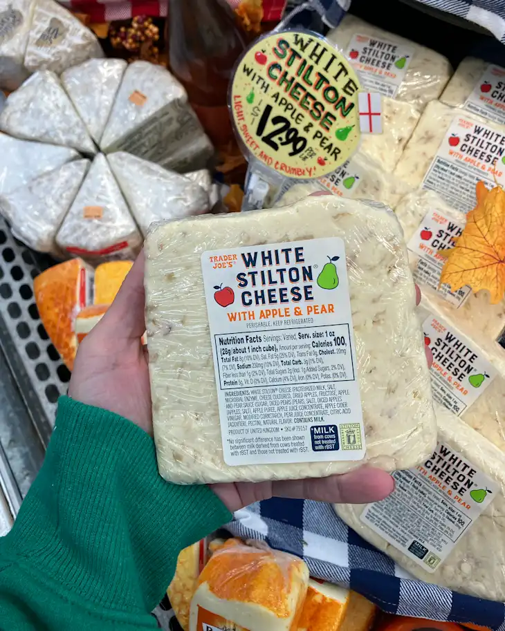 White Stilton cheese with apple and pear held in a hand, surrounded by other cheese packages and a price sign.