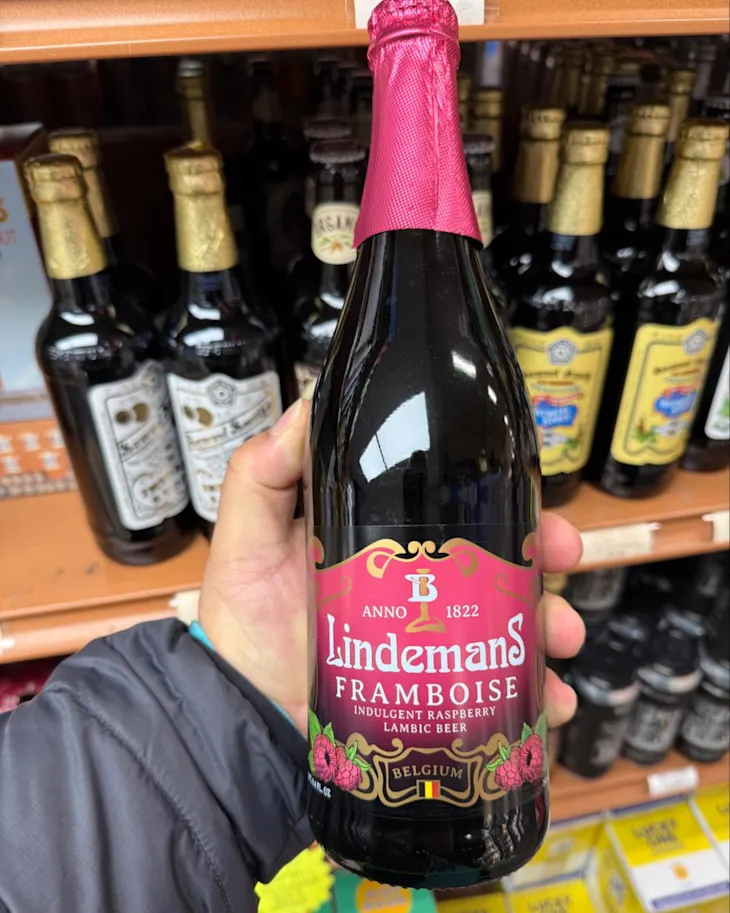 A person holding a bottle of Lindemans Framboise, a raspberry lambic beer, in a store aisle.